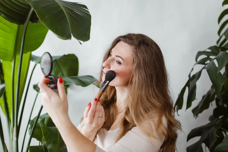 Woman holding a mirror and sweeping blush onto her cheek with a fluffy brush