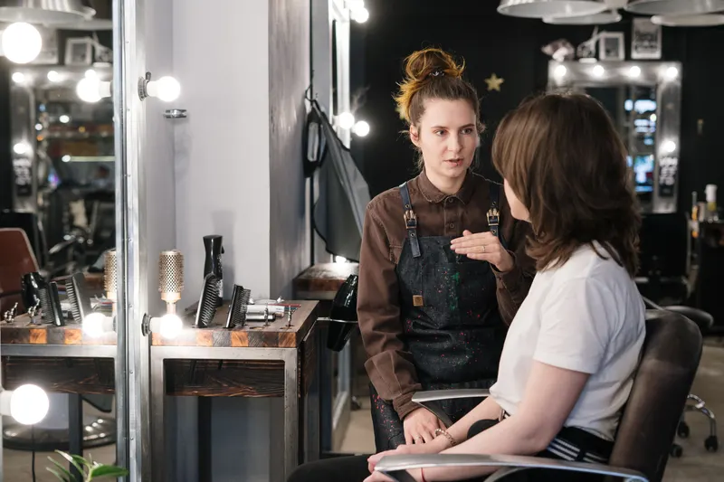 A stylist in an apron talks with a client in a salon chair during a consultation