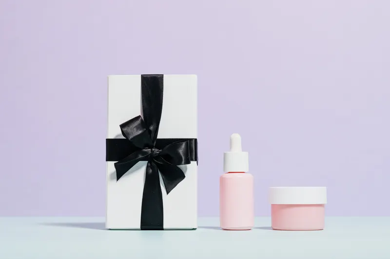 A wrapped gift box with a black ribbon beside a serum bottle and a jar of face cream