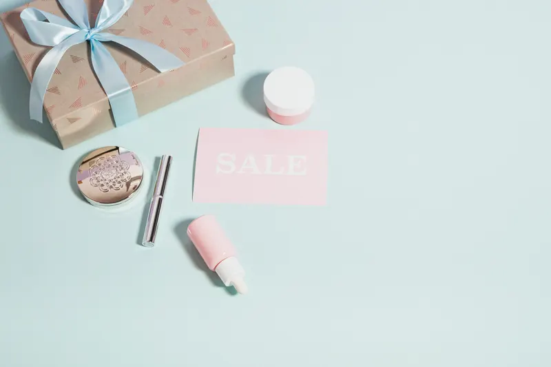 Makeup and skincare products beside a pink card that says sale on a pastel background