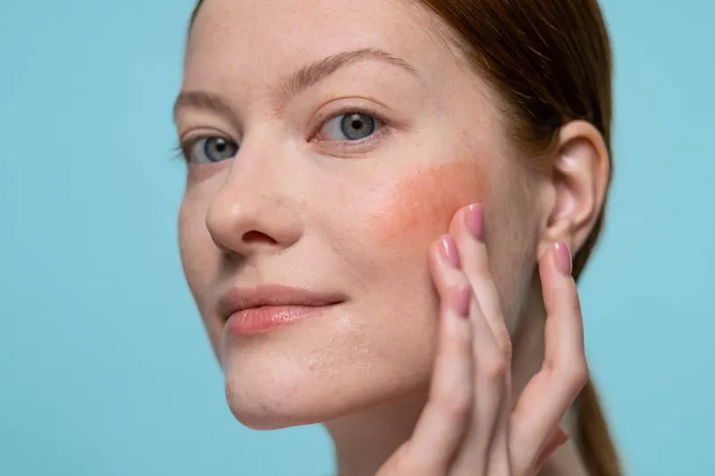 Woman with a soft peach blush on her cheek touching her face against a blue background