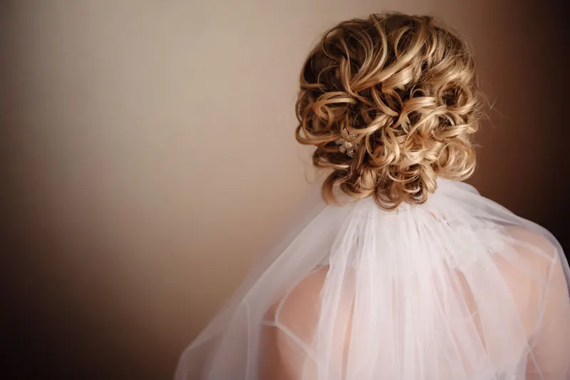 Back view of a curly blonde bridal updo with a sheer veil anchored beneath it