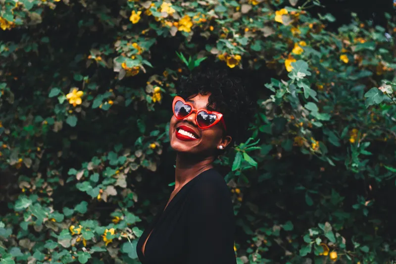 A laughing woman wearing a bright red lip and playful sunglasses in front of green leaves.