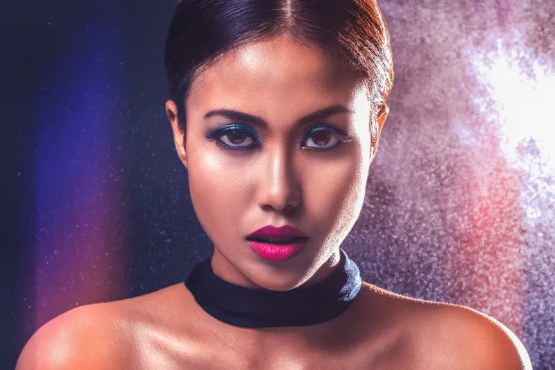 A woman with dark hair and luminous skin wears a vivid fuchsia lip against a dark background.