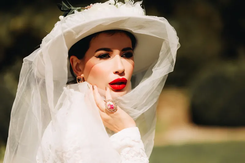 A bride in a sheer veil and wide-brimmed headpiece wears a classic blue-red lip.
