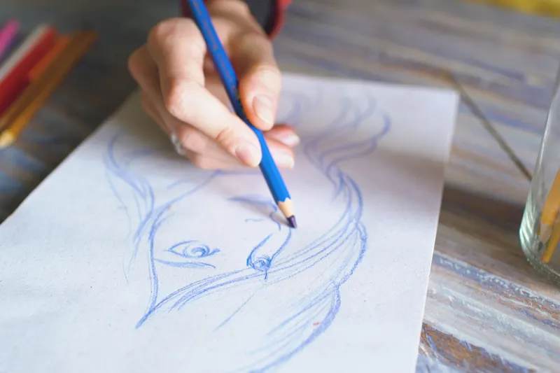 A hand sketches a face in blue colored pencil