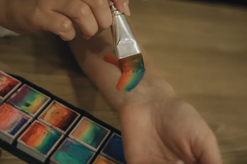 A brush paints rainbow stripes on an arm beside a palette of face paint cakes