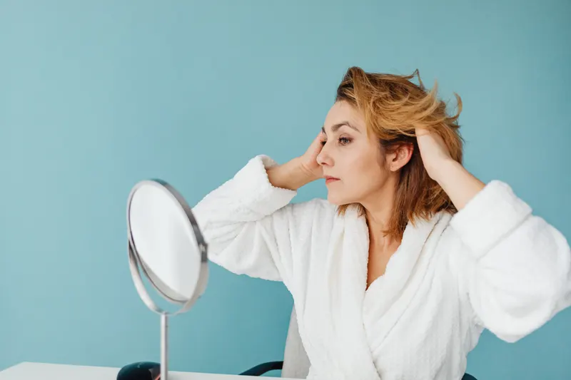 A woman in a bathrobe pulls her hair back from her face beside a small mirror
