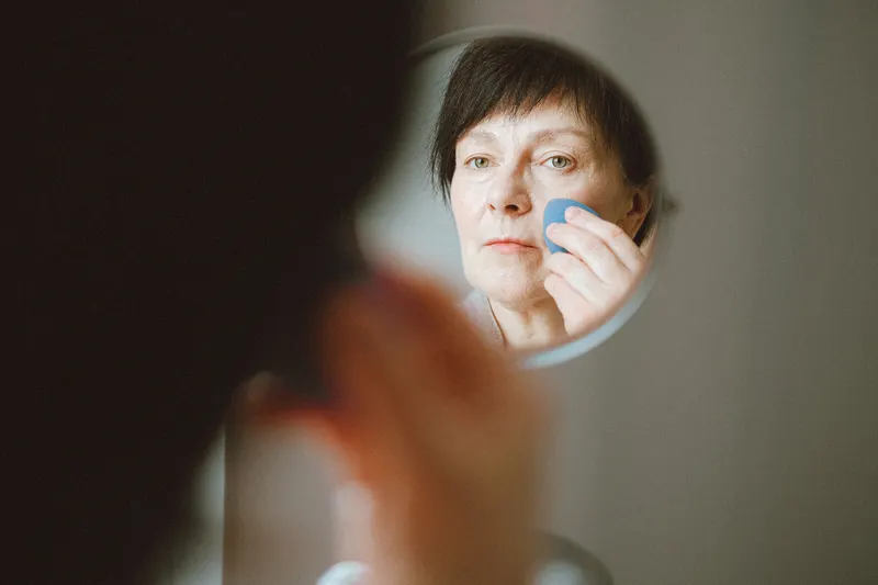 A woman with short dark hair blends foundation while checking a hand mirror