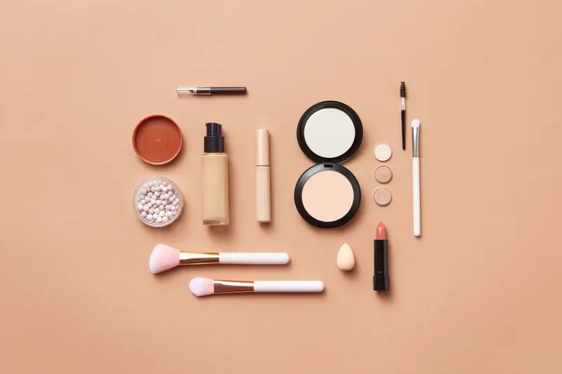 Flat lay of unbranded liquid foundation, pressed powders, a stick and brushes on a peach background