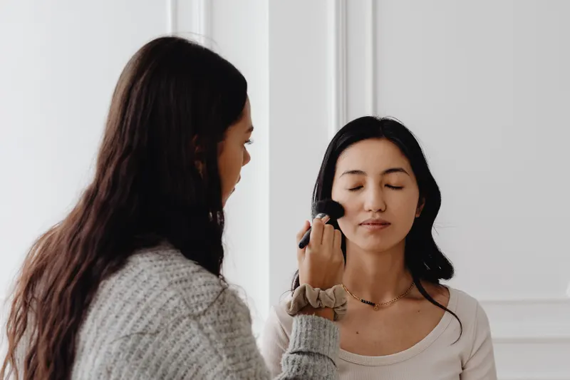 A makeup artist brushes foundation onto a client's cheek in a bright room lit by daylight, the kind of light a foundation shade finder suggests testing in.