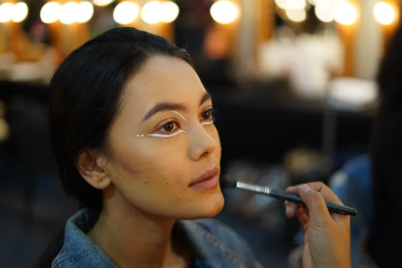 A makeup artist places lines of concealer under a client's eyes with a fine brush before blending.