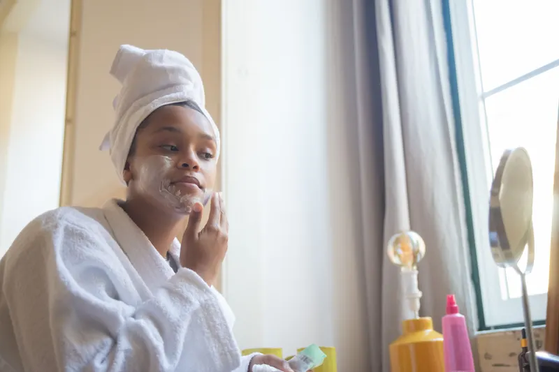 A woman in a bathrobe smooths face cream onto her cheek at home