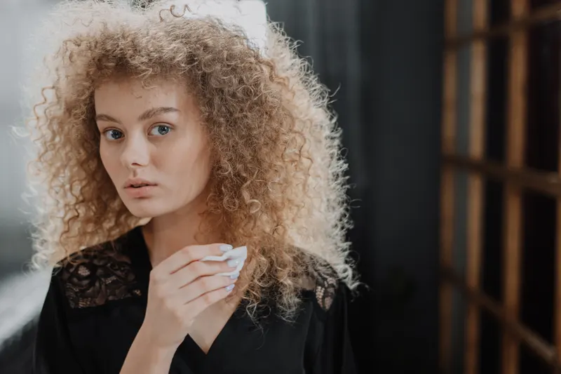 A woman with voluminous curly hair dabs her face with a cotton pad