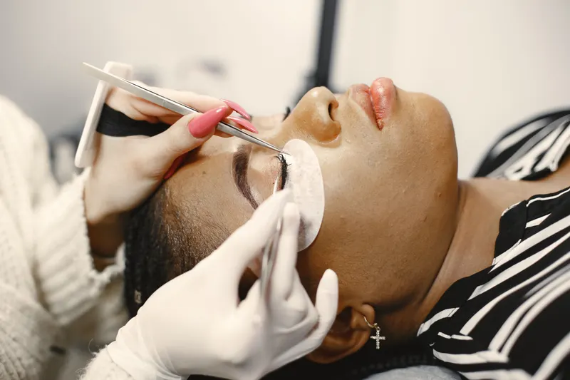 A lash technician uses tweezers to place lash extensions on a client lying back with an under-eye pad, the appointment a lash extension cost calculator budgets for.