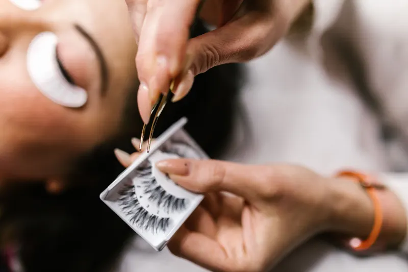 An artist lifts a pair of strip lashes from their tray with tweezers before applying them to a client.