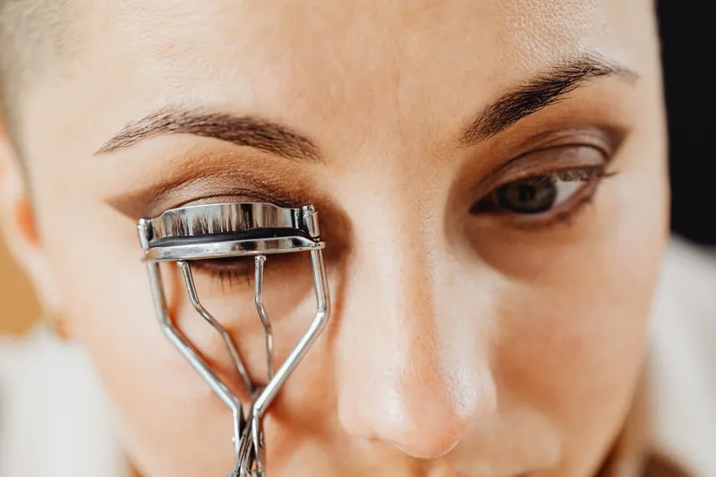A woman holds a metal eyelash curler at her lash line while doing her makeup.