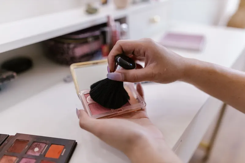 A makeup brush picks up a peach blush from an open compact on a white vanity.