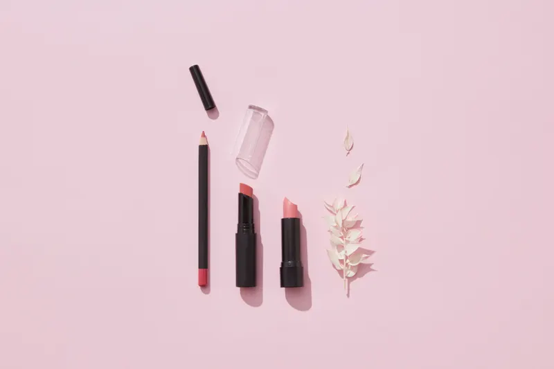 A soft pink lipstick and matching liner laid out on a pale pink background with a sprig of dried flowers.