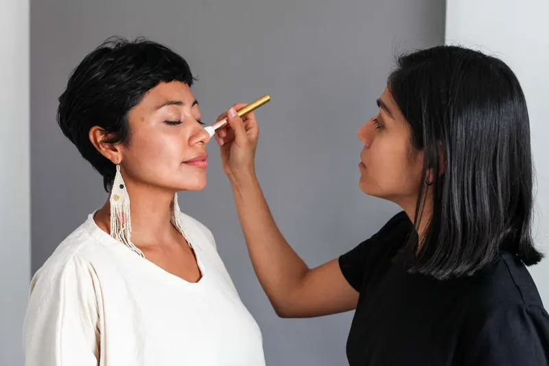 A makeup artist applies eye makeup to a woman with short dark hair sitting calmly with her eyes open.