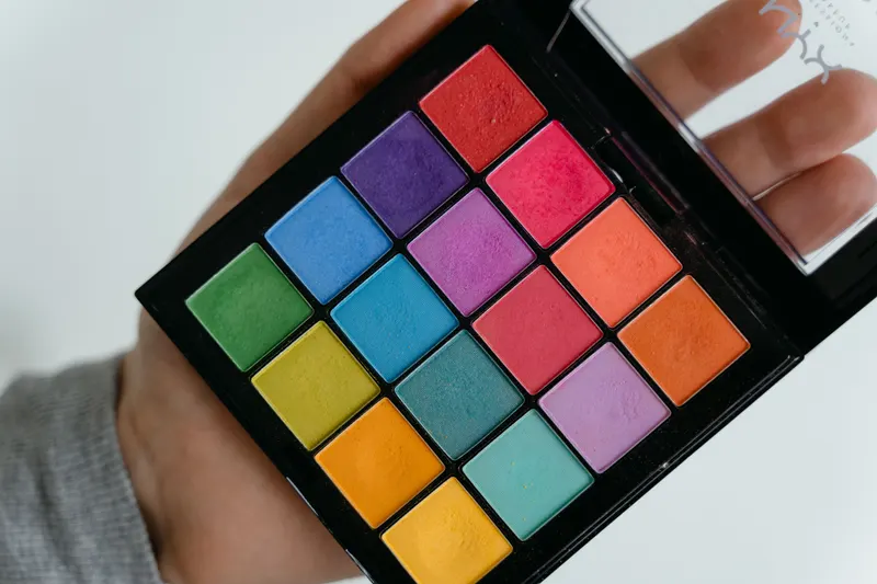 A hand holds a bright eyeshadow palette in a rainbow of colors