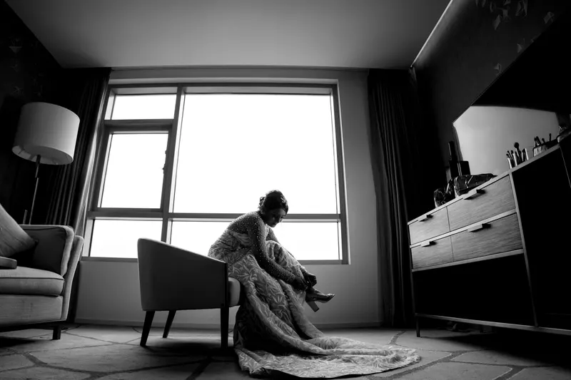 A bride sits in a hotel room beside a large bright window, in black and white