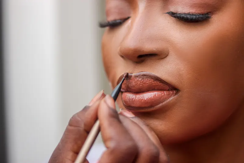 A makeup artist lines a bride's lips in a warm brown shade while she keeps her eyes closed.