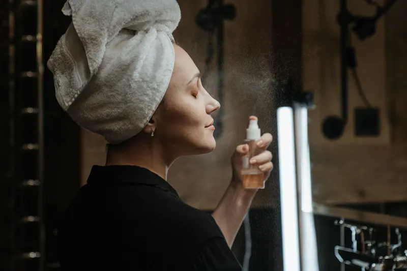 A woman with a towel on her hair mists her face with her eyes closed