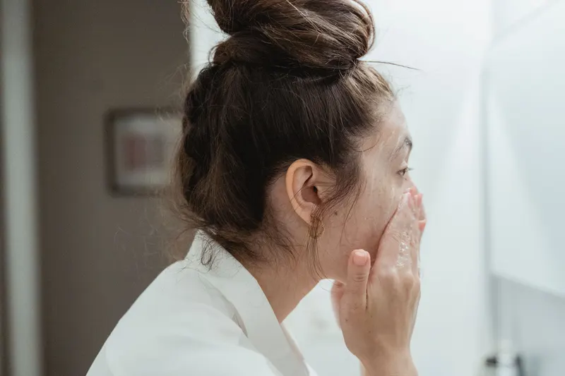 Woman with her hair in a bun washing her face with cleanser at the bathroom sink