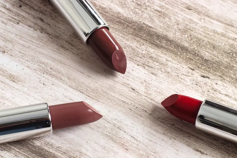 Two lipsticks in muted rose brown and warm mauve lie on pale wood.