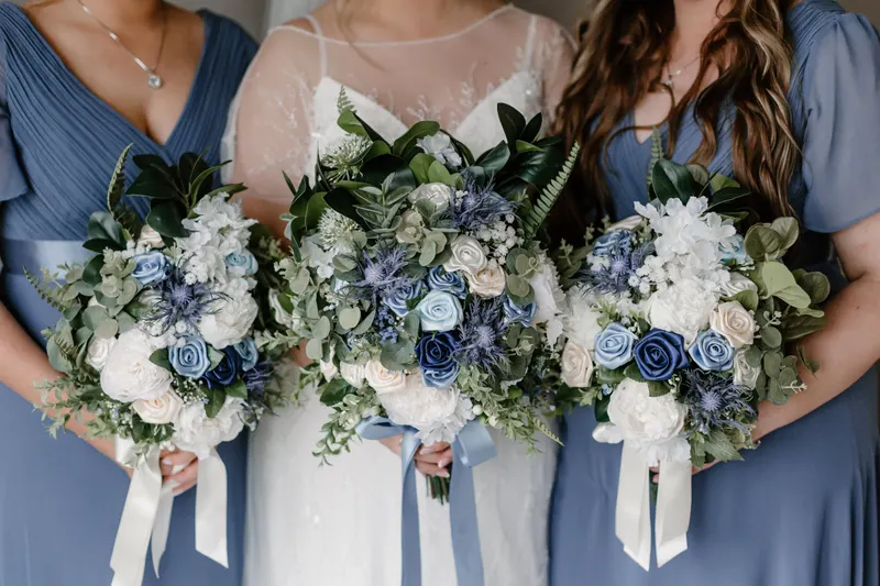 Bridesmaids in dusty blue dresses hold bouquets of white and slate blue flowers beside a bride, the muted cool tones of a soft summer color palette.