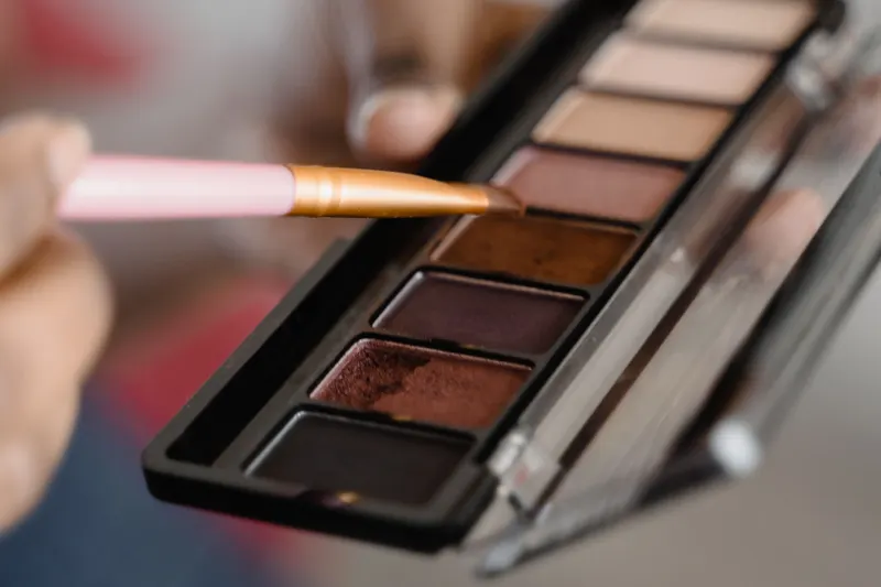 A brush dips into a palette of bronze, copper and warm brown eyeshadows.