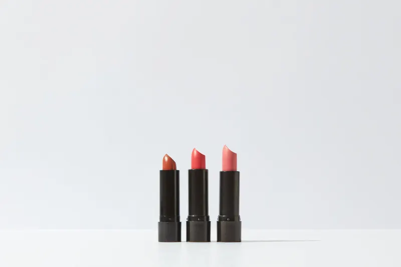 Three lipsticks in coral, warm pink and peach stand in a row on a white surface.