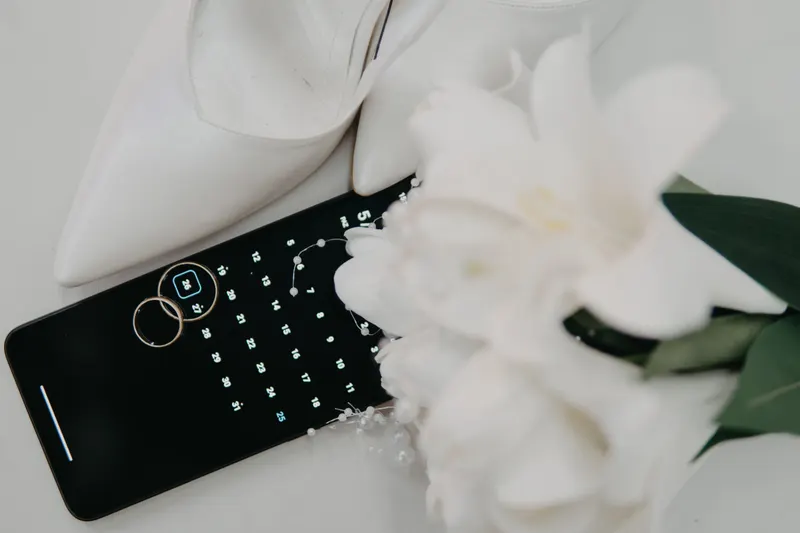 Two wedding rings rest on a phone calendar beside white bridal shoes and a bouquet, a reminder to plan the beauty steps before the wedding.