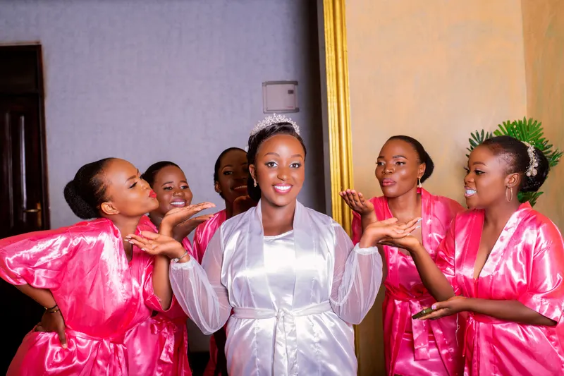A bride in a white robe celebrates with five bridesmaids in pink robes while they get ready for the wedding.