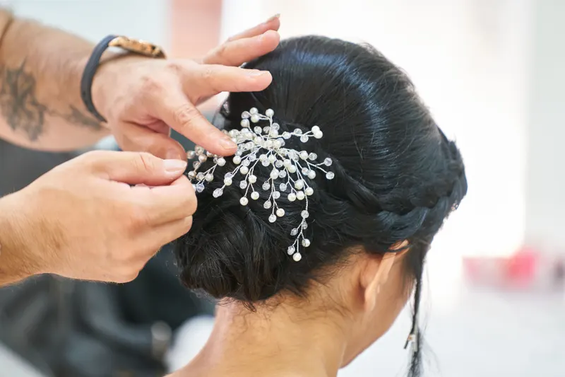 A hair stylist pins a crystal hairpiece into a dark braided bridal updo.