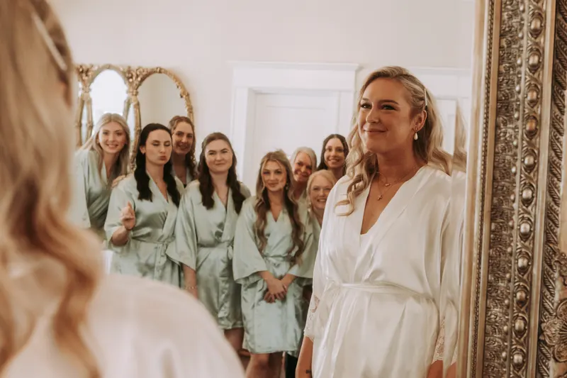 A bride in a white robe smiles into a gold mirror while a large bridal party in matching sage robes waits behind her on the wedding morning.
