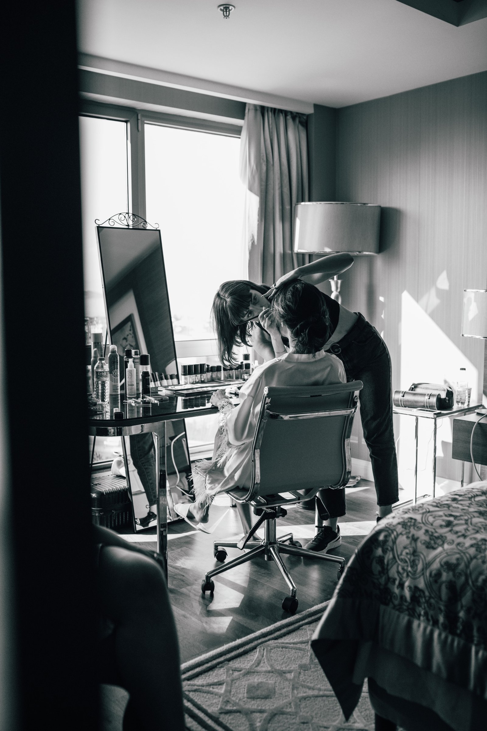 A professional stylist provides on-location wedding hair and makeup services in a bright, softly lit getting-ready space.