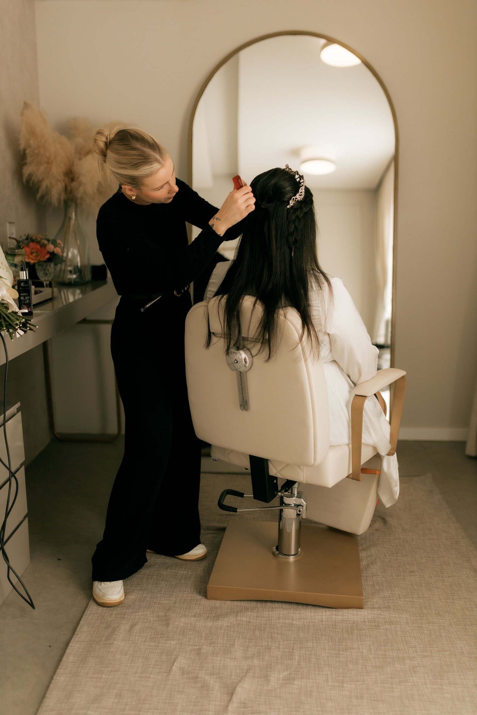 A wedding hair stylist works on a bride's updo at a vanity mirror before the ceremony.