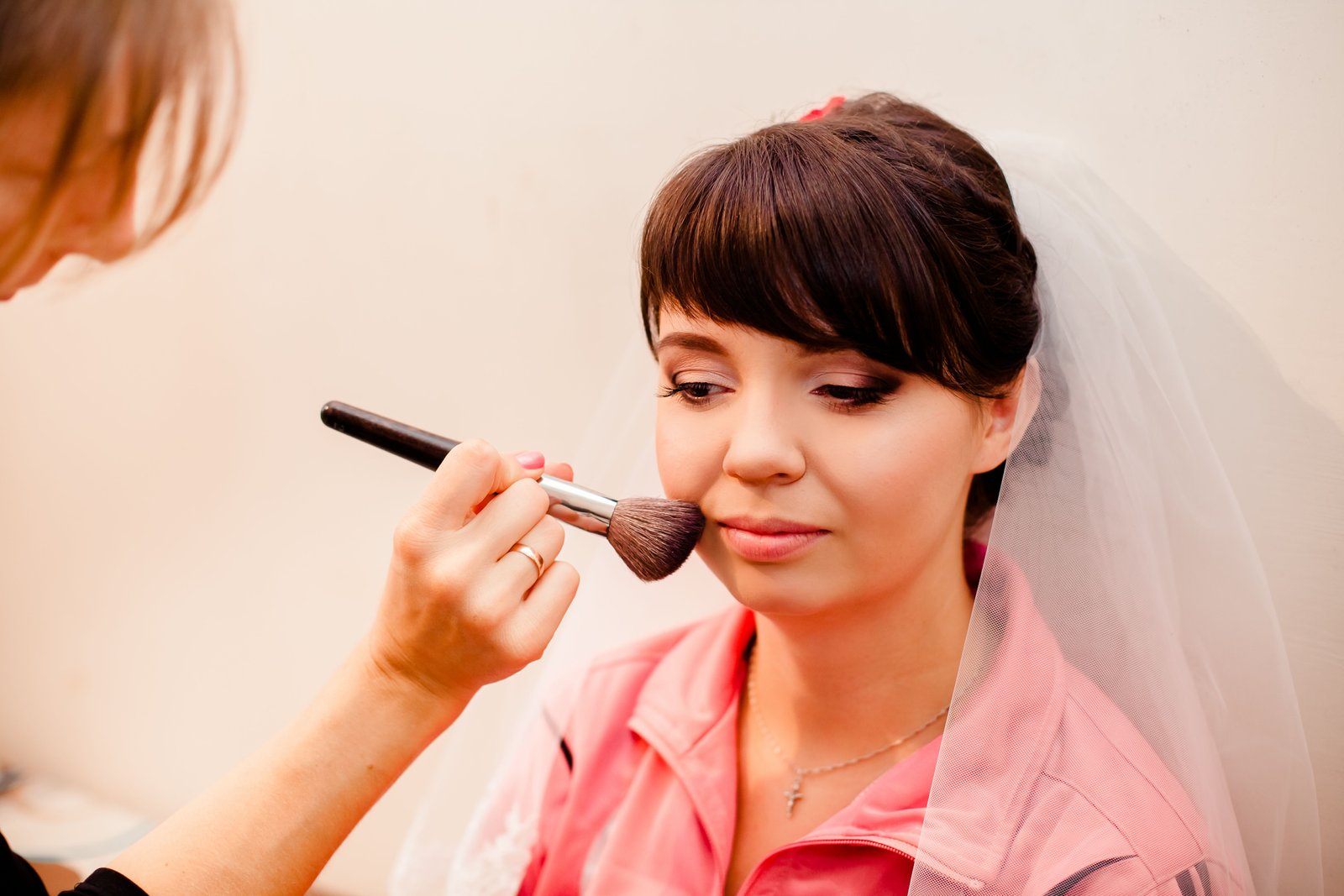 A makeup artist applies blush to a bride wearing a veil, part of a full bridal makeup application.