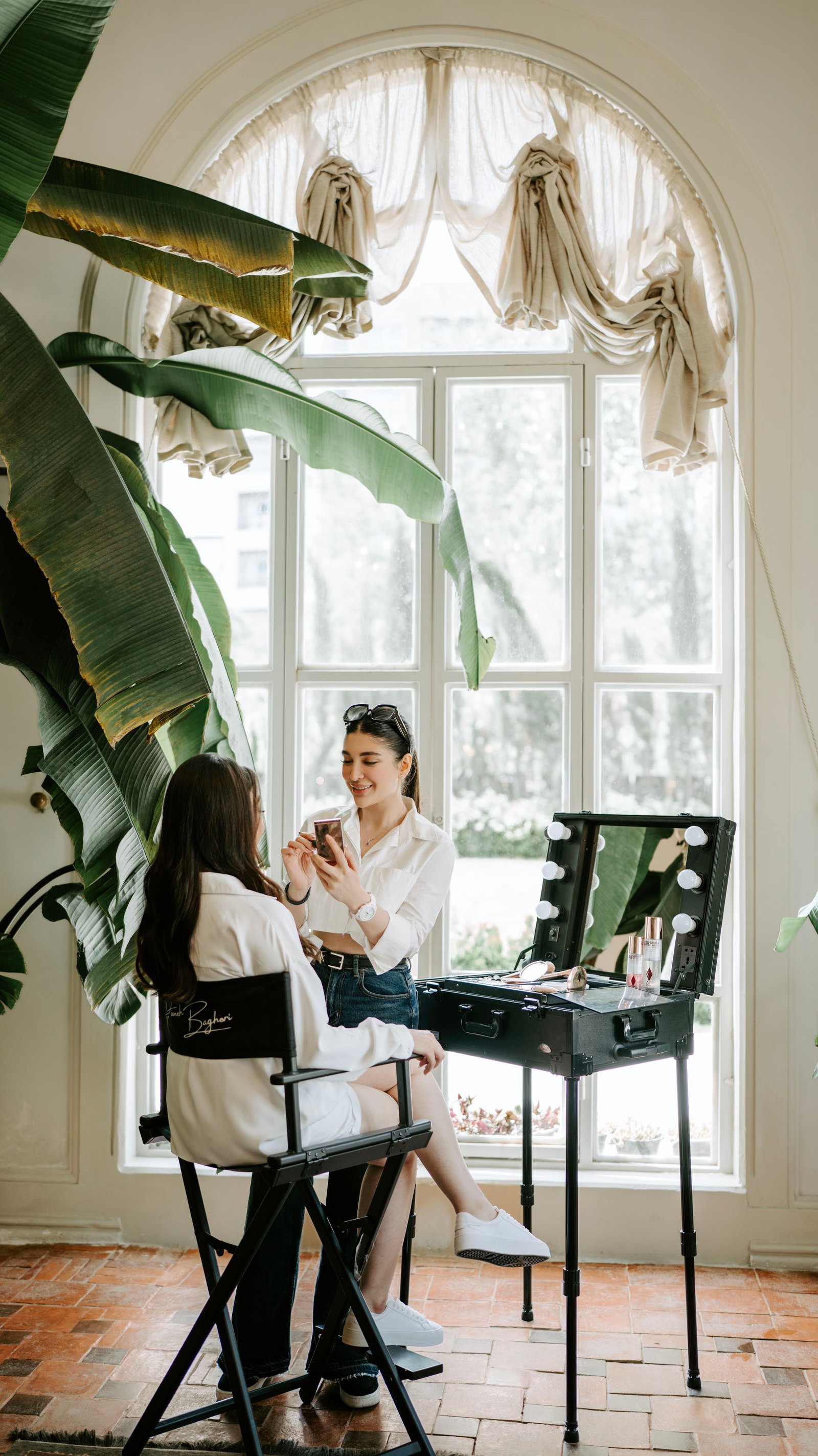 A makeup artist applies cosmetics to a bride in a sunlit room, illustrating what wedding hair and makeup packages include.