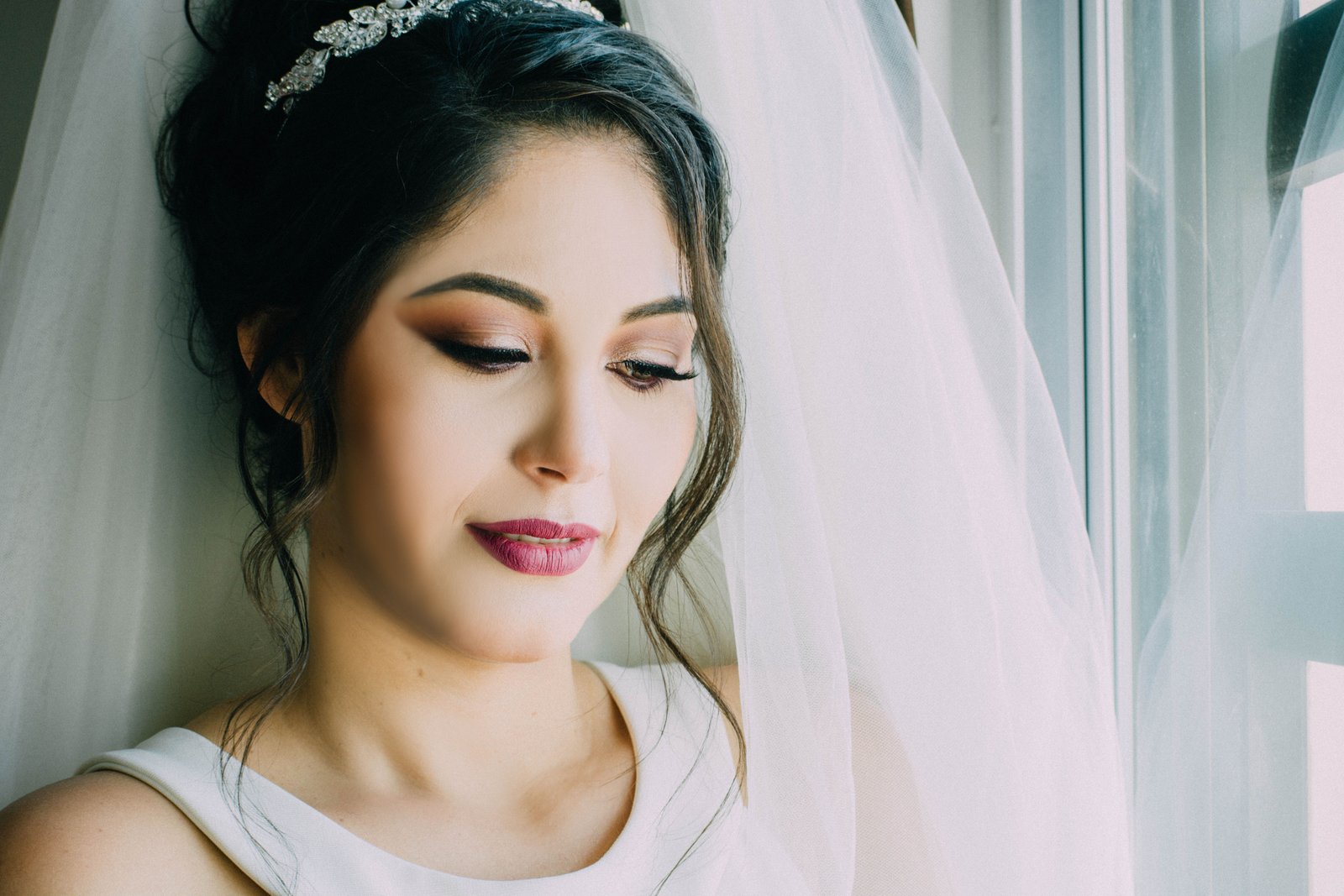 A bride with finished bridal makeup and a veil looks down softly by a window.