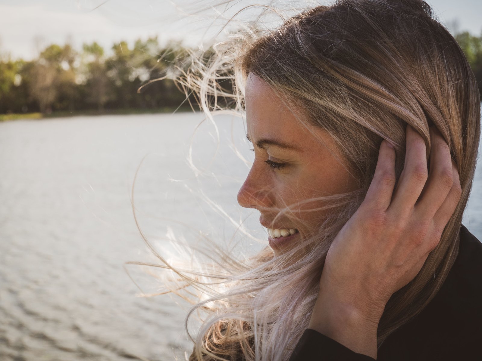 Woman with blonde hair smiling by a breezy lakeside